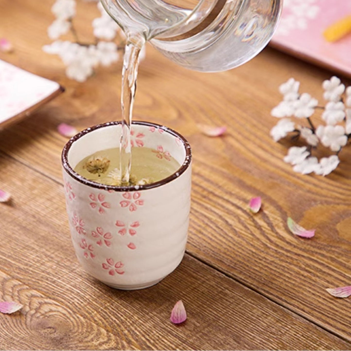Tea being poured into a decorative teacup on a wooden table with flowers and snacks.