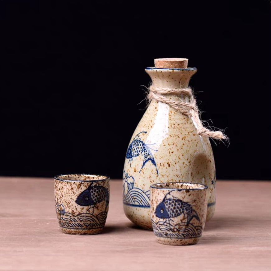 A person holding a ceramic sake bottle with blue fish patterns, alongside matching ceramic cups arranged on a table with a plant in the background.