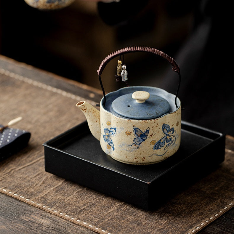A ceramic teapot with blue butterfly illustrations and a dark metal handle, resting on a black tray on a wooden table.