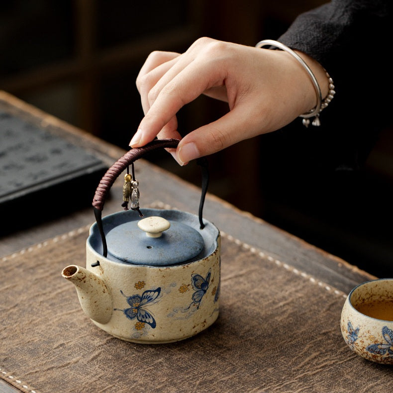 A ceramic tea kettle with a blue lid and blue butterfly motifs on a cream body, being held by a person's hand over a wooden table. A small matching cup is partially visible on the right.