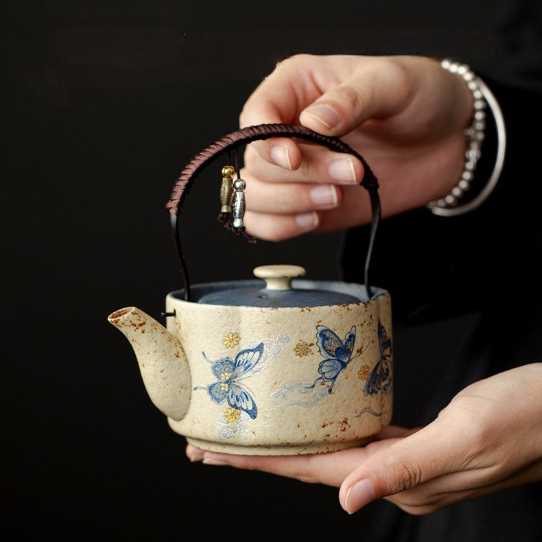 A ceramic tea kettle with a cream-colored glaze, blue butterfly illustrations, and a dark brown woven handle grip, held by two hands against a dark background.