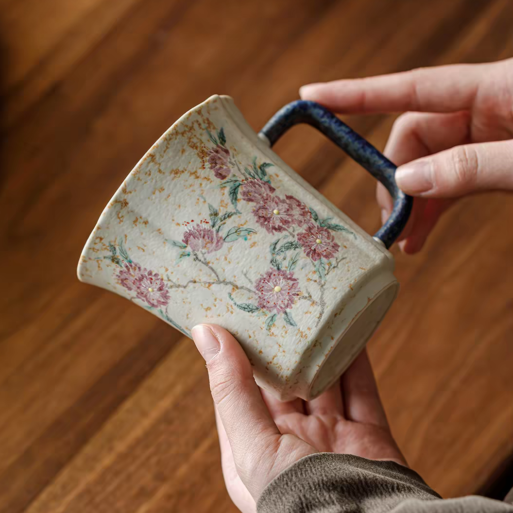 A handmade ceramic mug with a floral pattern and a dark blue handle, held by hands over a wooden table background.