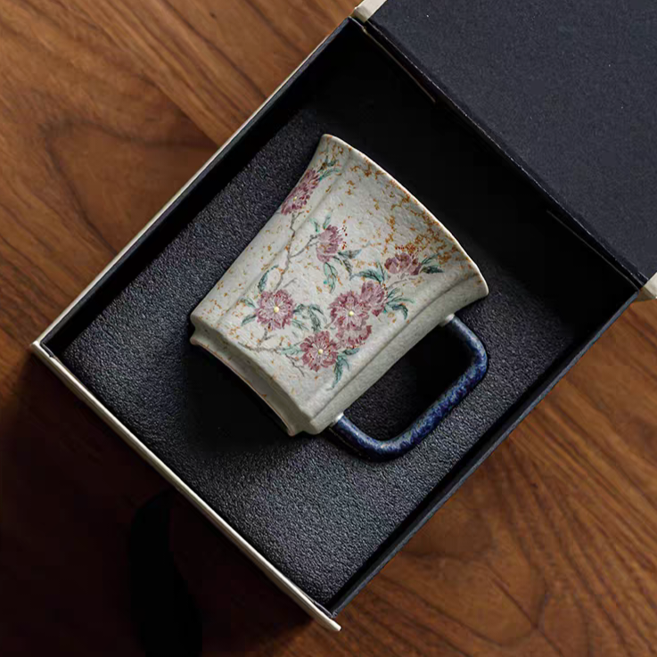 A small ceramic mug with a floral pattern and a dark blue handle, presented inside an open black gift box on a wooden surface.