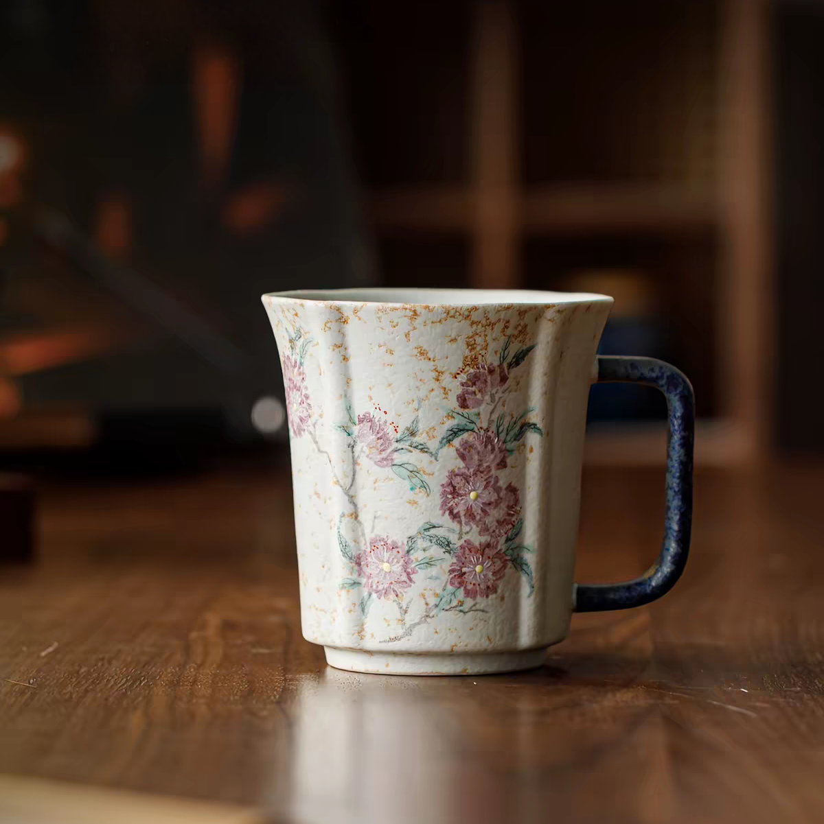 A vintage-style ceramic mug with a distressed cream body, pink floral pattern, and a dark blue handle, resting on a wooden table.