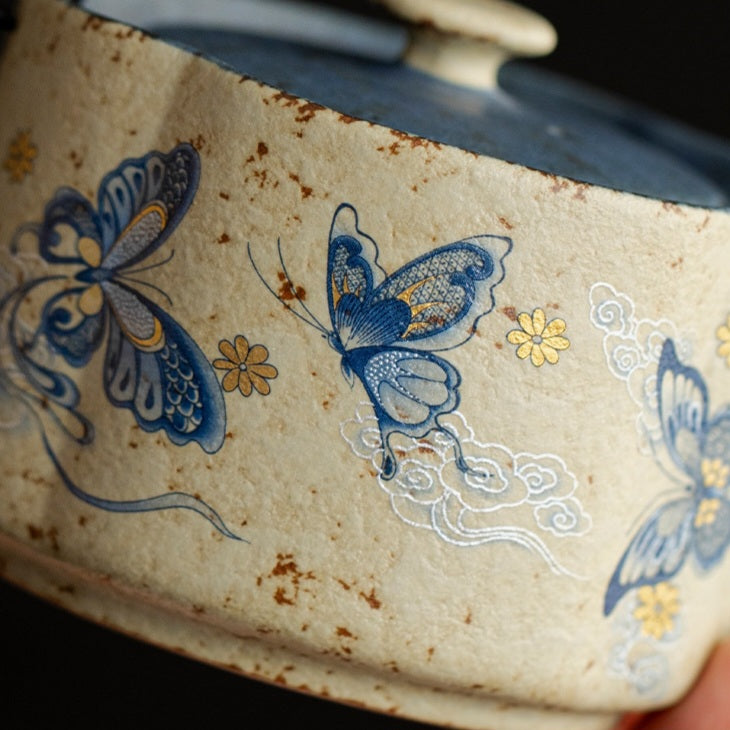 Close-up of a ceramic container or bowl with a distressed cream glaze, decorated with blue butterflies, small yellow flowers, and white cloud motifs.
