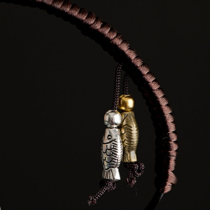 A close-up of a dark brown braided cord necklace with two hanging charms: one silver fish-shaped pendant and one gold fish-shaped pendant, set against a solid black background.