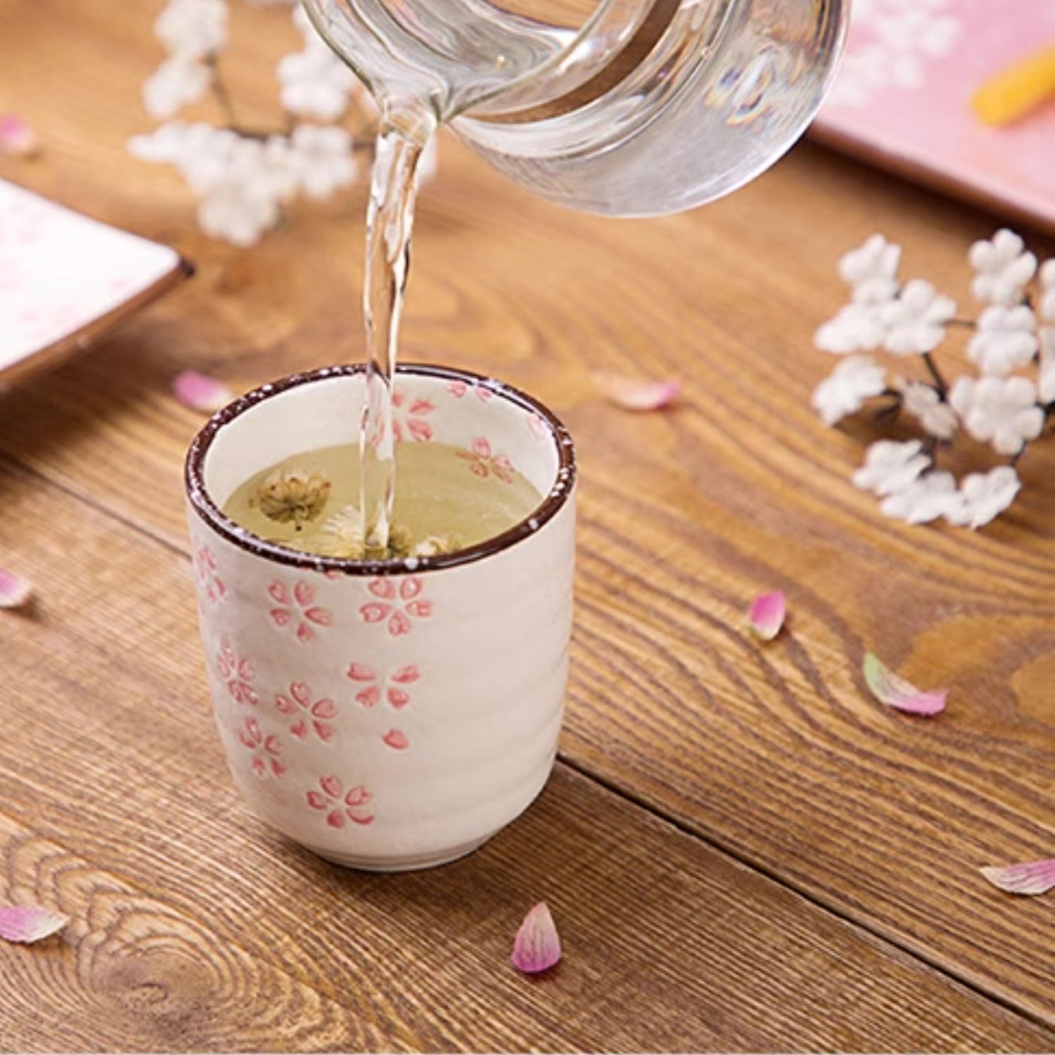 Tea being poured into a decorative teacup on a wooden table with flowers and snacks.